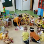 Dzieci ubranie w żółte bluzeczki z żółtymi opaskami na włosach siedzą ustawione w koło. Wszyscy patrzą na nauczycielkę, która rysuje na białej kartce położonej na podłodze.