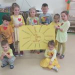 Sześcioro dzieci trzyma wykonaną przez siebie żółtą planszę ze słoneczkiem i promyczkami. Dwoje dzieci siedzi na podłodze.