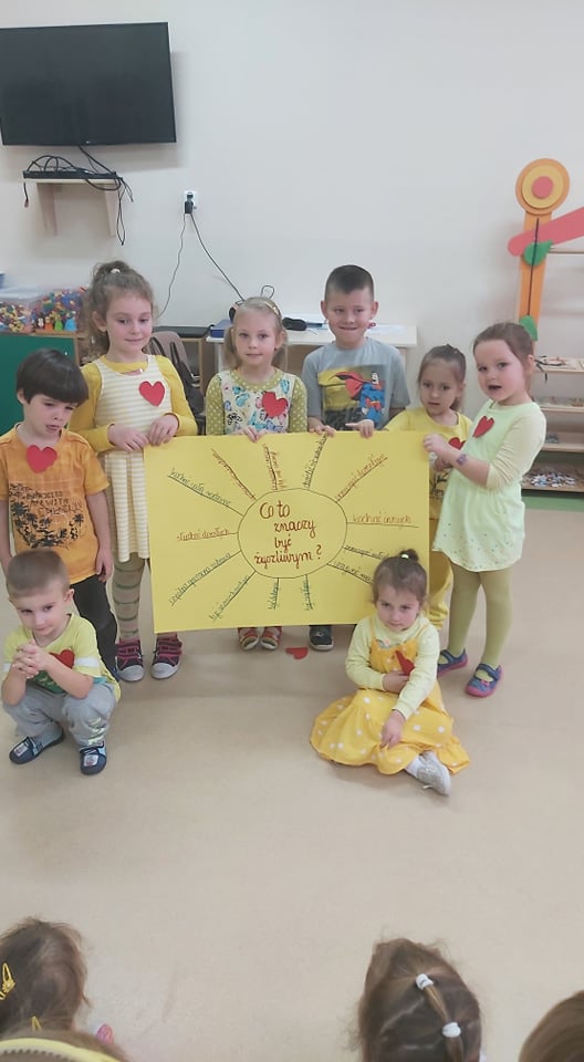 Sześcioro dzieci trzyma wykonaną przez siebie żółtą planszę ze słoneczkiem i promyczkami. Dwoje dzieci siedzi na podłodze.
