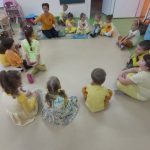 Dzieci w żółtych ubraniach siedzą po turecku na podłodze tworząc kółko. Słuchają uważnie nauczycielki.