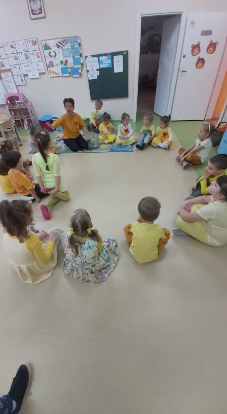 Dzieci w żółtych ubraniach siedzą po turecku na podłodze tworząc kółko. Słuchają uważnie nauczycielki.