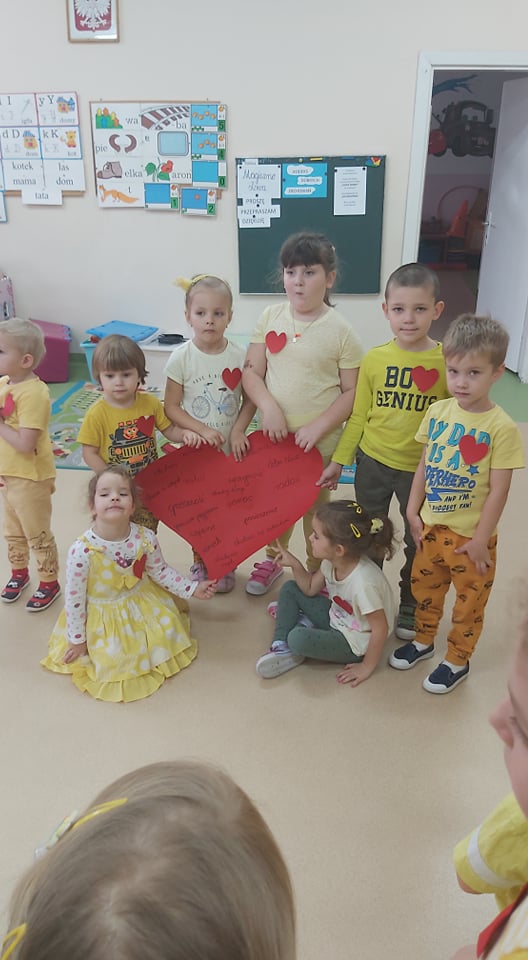 Ośmiro dzieci trzyma w rękach ogromne papierowe czerwone serce.