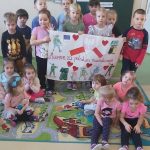 Grupa dzieci. Dziesięć dzieci stoi a siedmioro siedzi na kolorowym dywanie. Dwoje dzieci trzyma w rękach przygotowaną przez siebie planszę.