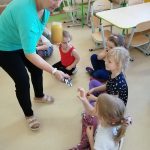 Nauczycielka częstuje dzieci siedzące w półkolu na podłodze miodem.