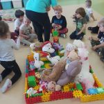 Na pierwszym planie maskotki misiów ułożone na platformie z klocków. W tle dzieci siedzace w półkolu i nauczycielka rozdająca dzieciom miód