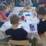 Dzieci siedzą dookoła stołu nakrytego białą folią wykonują prace plastyczne farbami. Na stole leża farbki, słoiczki z wodą pędzle oraz wykonywane przez dzieci prace.