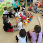 Dzieci siedzą po turecku na podłodze. Na środku grupa maskotek ułożona na platformie z klocków. Dzieci słuchają opowiadania nauczycielki.