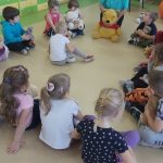 Dzieci ustawione w półkole siedzą na podłodze. Nauczycielka w niebieskiej bluzie trzyma przed sobą maskotkę Kubusia Puchatka.