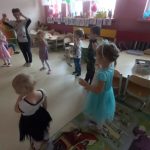 Dzieci wraz z nauczycielką ustawione w półkole, ubrane w kolorowe stroje tańczą kaczuchy.