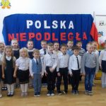 Dzieci w strojach galowych stoja dwuszeregu na tle napisu Polska Niepodległa