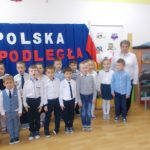 Dzieci w strojach galowych stoja dwuszeregu na tle napisu Polska Niepodległa
