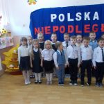 Dzieci w strojach galowych stoja dwuszeregu na tle napisu Polska Niepodległa