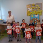 Dzieci ubrane w białe bluzeczki ustawione dwuszeregu trzymają w rękach wykonana przez siebie godło Polski. Za nimi stoi nauczycielka.