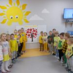 Dzieci w żółtych strojach stoją w formie promieni. Na ścianie przypięta praca plastyczna wykonana przez dzieci: brązowe drzewo z czerwonymi sercami zamiast liści. Wysoko na ścianie żółte słoneczko.