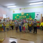 Duża grupa dzieci ubranych na zółto wraz z nauczycielką stoi na tle zielonej kotary z napisem dzień życzliwości.