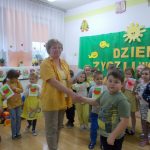 Grupa dzieci stoi na tle zielonej kotary z napisem dzień życzliwości. Na pierwszym planie nauczycielka w żółtej koszulce trzyma za rękę chłopca.