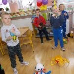 Czworo dzieci w kolorowych ubraniach stoi luźno. Przed nimi na podłodze leżą maskotki. W tle udekorowane firanką i kolorowymi balonami okno..