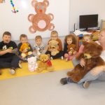 grupa dzieci ustawiona w półkole, siedzi po turecku trzymając w rękach misie. W tle widać plakat z dużym brązowym misiem