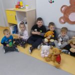 pięcioro dzieci siedzi po turecku. na pierwszym planie siedzą: bały i brązowy miś. W tle plakat z brązowym misiem i szafka z zabawkami dla dzieci.