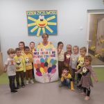 Dzieci w żółtych strojach wraz z nauczycielką stoją w półkolu.Nauczycielka trzyma pracę plstyczną wykonana przez dzieci. W tle na ścianie słoneczko z promykami.