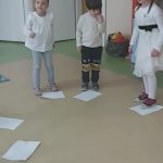 Dzieci w białych strojach stoją po okręgu. Przed nimi leża białe kartki papieru.