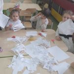 Gruba ubranych na biało dzieci siedzi przy stokuku wycinając z białego papieru gwiazdy.