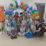Grupa dzieci ubranych w stroje karnawałowe wraz z opiekunami pozuje pod łukiem z kolorowych balonów.