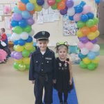 Chłopczyk w stroju policjanta oraz dziewczynka w czarnej sukience stoją na granatowym dywanie. W tle łuk z kolorowych balonów.