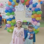 Dwie dziewczynki w strojach karnawałowych pozują do zdjęcia na tle łuku z kolorowych balonów.