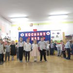 Dzieci ustawione w dwóch rzędach na tle napisu kochamy was recytują wiersze.