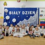 Duża grupa dzieci siedzi po turecku. W tle biały napis na niebiskim materiale: biały dzień