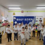 Duża grupa dzieci ubranych na biało stoi a tle napisu biały dzień trzymając w rękach białe kartki.