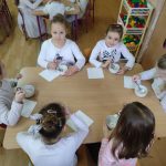 sześcioro dzieci ubranych na biało siedzi dookoła stoika. Przed nimi białe kartki i pojemniki. Dzieci tworzą pracę plastyczną.
