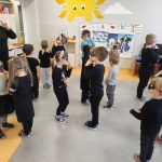 Zdjęcie przedstawia grupę dzieci ubranych na szaro i czarno z maseczkami kota na twarzy. Są w trakcie zabawy. Stoją w parach, twarzami do siebie, ich dłonie skierowane są w kierunku nosów. Z boku stoi nauczycielka w czarnym stroju. Pokazuje dzieciom, jaki gest mają wykonać.