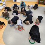Grupa dzieci w czarno - szarych strojach podczas zabawy o kotkach. Znajdują się w sali zabaw grupy Promyczków. Klęczą pochyleni w kierunku plastikowych obręczy - misek.