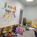 Sala grupy Pszczółek. Grupa dzieci w kolorowych strojach i kartonowych czapkach - kredkach siedzi oparta o szafki. Dzieci zwrócone twarzą w kierunku nauczycielki, która siedzi z boku. W dłoni trzyma kartkę. W tle kolorowe szafki, misie i duże klocki.