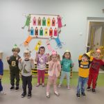 Grupa 8 dzieci w kolorowych strojach. Na głowach mają kartonowe czapeczki w formie stożka - podstruganej kredki. Ręce mają podniesione lekko w górę. W tle znajduje się tablica z kartonowymi kredkami, na nich napis Dzień Kredki oraz kartonowe kolorowe ludziki.