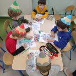 Pięcioro dzieci w kartonowych czapkach - kredkach siedzi przy stolikach kolorując obrazki. Na stoliku znajdują się też przybory plastyczne - kredki, pisaki, nożyczki.
