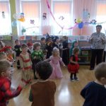 Zdjęcie przedstawia grupę dzieci oraz nauczycielkę. Bawią się oni na zabawie karnawałowej. Dzieci mają stroje z bajkowego przebrania. Część dzieci tworzy małe koło, część stoi obok okien, na zewnątrz koła. Sala udekorowana balonami, serpentynami, wycinankami bałwanów i sów.