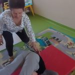 Chłopiec w szarych spodniach i ciemnogranatowej bluzie wykonuje na czerwonym materacu przewrót w tył Celina Asekuruje go wychowawczyni