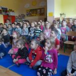 Zdjęcie przedstawia salę zabaw grupy Mądrych Sów. W sali znajduje się grupa kilkudziesięciu przedszkolaków: część z nich siedzi na krzesełkach ( w środku i w tyle), część na niebieskich matach z przodu. Z boku, po lewej stronie siedzi nauczycielka. Dzieci wpatrują się w coś, co znajduje się przed nimi.