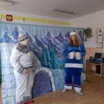 Dwie aktorki odgrywają scenkę w przedstawieniu. Jedna z nich ubrana w dwuczęściowy biały strój z szarymi wzorami i białą czapkę we wzory, druga w dwuczęściowy strój niebieski z białymi obszyciami i białą czapkę z nausznikami. Za nimi znajduje się rozłożony stelarz z tłem do przedstawienia: wejście do igloo, z boku po prawej stronie rosną ośnieżone świerki, w tyle widoczne są ośnieżone szczyty gór.