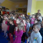 Na zdjęciu znajduje się grupa około 30 stojących przedszkolaków. Dzieci biorą udział w proponowanej zabawie. Ich twarze są zwrócone w jednym kierunku, większość ma dłoń przyłożoną do czoła w geście patrzenia. Po prawej stronie na końcu stoją dwie nauczycielki, po lewej z boku opiekunka dzieci.