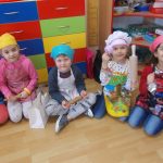 Na tle kolorowych szafek przedszkolnych siedzi pięcioro dzieci - dwoje w siadzie skrzyżnym, troje w siadzie klęcznym. Pierwsze z dzieci przedstawionych na zdjęciu ujęte jest tylko fragmentarycznie, nie widać jego głowy. Dzieci mają na sobie kolorowe stroje, zakryte krótkimi kuchennymi fartuszkami, ich włosy są osłonięte chusteczkami w kolorach: żółtym, niebieskim, białym i różowym. W rękach trzymają wałki kuchenne oraz foremki.