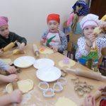 Przy stoliku na jadalni siedzi grupa przedszkolaków: troje dzieci widocznych w całości i troje fragmentarycznie. Dzieci nakryły głowy kolorowymi chusteczkami, założyły też fartuchy kuchenne. Na stoliku przed nimi znajdują się trzy białe talerze zawierające mąkę, cukier i białko z jajek. Leżą też wałki i foremki.. Dzieci wałkują małe kawałki ciasta na podsypanym mąką stole. Jedna z dziewczynek pokazuje wycięty przez siebie kształt ciastka - okrągły kwiatek