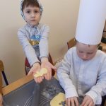 Zdjęcie przedstawia dwóch chłopców. Jeden z nich stoi w chusteczce na głowie, wyciąga przed siebie złączone dłonie - i pokazuje wycięty kształt ludzika.. Drugi chłopiec siedzi z pochyloną głową. Ma na sobie białą koszulę i białe, kartonowe nakrycie głowy. Na rozwałkowanym przed sobą kawałku ciasta wycina foremką kształt ciastka. W tle widoczny fragment innego stolika oraz dłonie dziecka wałkującego ciasto.