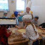 Zdjęcie zrobione w jadalni. Widać na nim dzieci ubrane w kolorowe stroje, fartuszki kuchenne i nakrycia głowy. Pracują one przy stolikach wałkując ciasto, wycinając je foremkami. Z lewej strony widoczna wyciągnięta czarna blaszka wyłożona papierem, na którą dzieci wykładają swoje ciastka do upieczenia.
