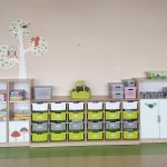szafki przedszkolne w kolorach szaro - zielono - białym. Na szafkach stoją pojemniki w tych samych kolorach. Na półeczkach leża książki, puzzle . Na ścianie przyczepione wycięte ze sklejki drzewo z naklejonym obrazkiem sowy.