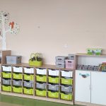 szafki przedszkolne w kolorach szaro - zielono - białym. Na szafkach stoją pojemniki w tych samych kolorach. Na półeczkach leża książki, puzzle . Na ścianie przyczepione wycięte ze sklejki drzewo z naklejonym obrazkiem sowy.