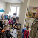 Dzieci siedzą w czytelni bibliotecznej. Z prawej strony stoi zwrócony do nich twarzą mężczyzna, ubrany w jasną kurtkę. Jest to Pan Stanisław Majcher z Jadownik Mokrych, malarz, autor prac wystawianych w bibliotece. Za nim widoczna jest biała szafka. Stoją na niej obrazy z motywami kwiatów. Z prawej strony widać siedzącą nauczycielkę. W głębi na ścianie widoczny fragment napisu ,, czytelnia'' - i pomalowane trzy niebieskie, poziome pasy. Po prawej stronie w głębi sali widać okno biblioteczne.